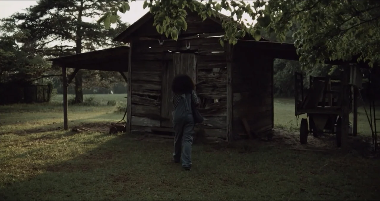 Eye for an Eye (2025) movie still: fullbody — An aged, dilapidated wooden shed sits in a grassy clearing surrounded by large trees. A woman in denim…