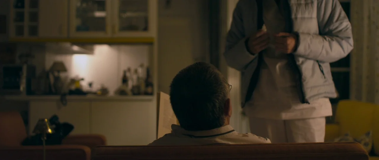 Alps (2011) movie still: medium — A dimly lit room with a man sitting on a sofa reading a book. A person wearing a puffer jacket stands facing him…