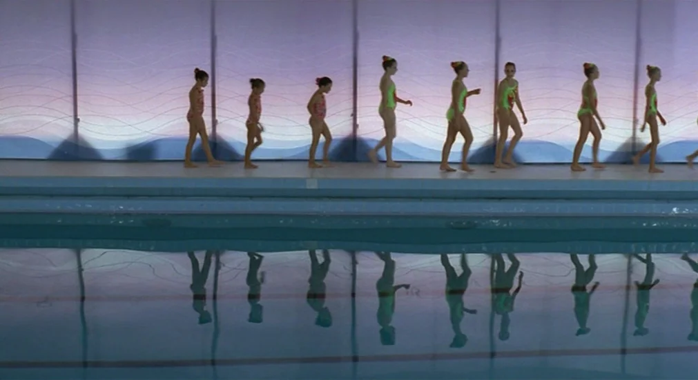 Water Lillies (2007) movie still: wide — A line of children in matching swimsuits walks along the edge of an indoor pool. Their movements and bodies…