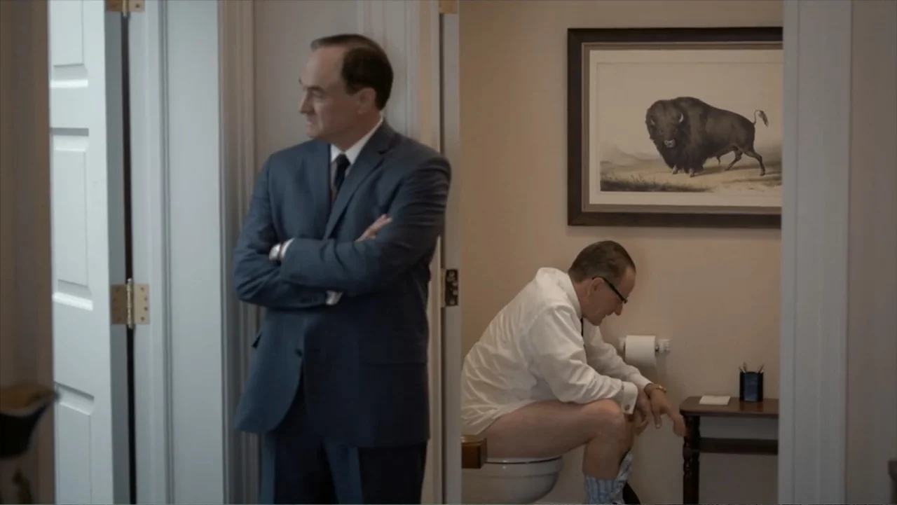 All the Way (2016) movie still: medium — A sterile bathroom with beige walls contains a framed print of a buffalo. One man wearing a business suit…