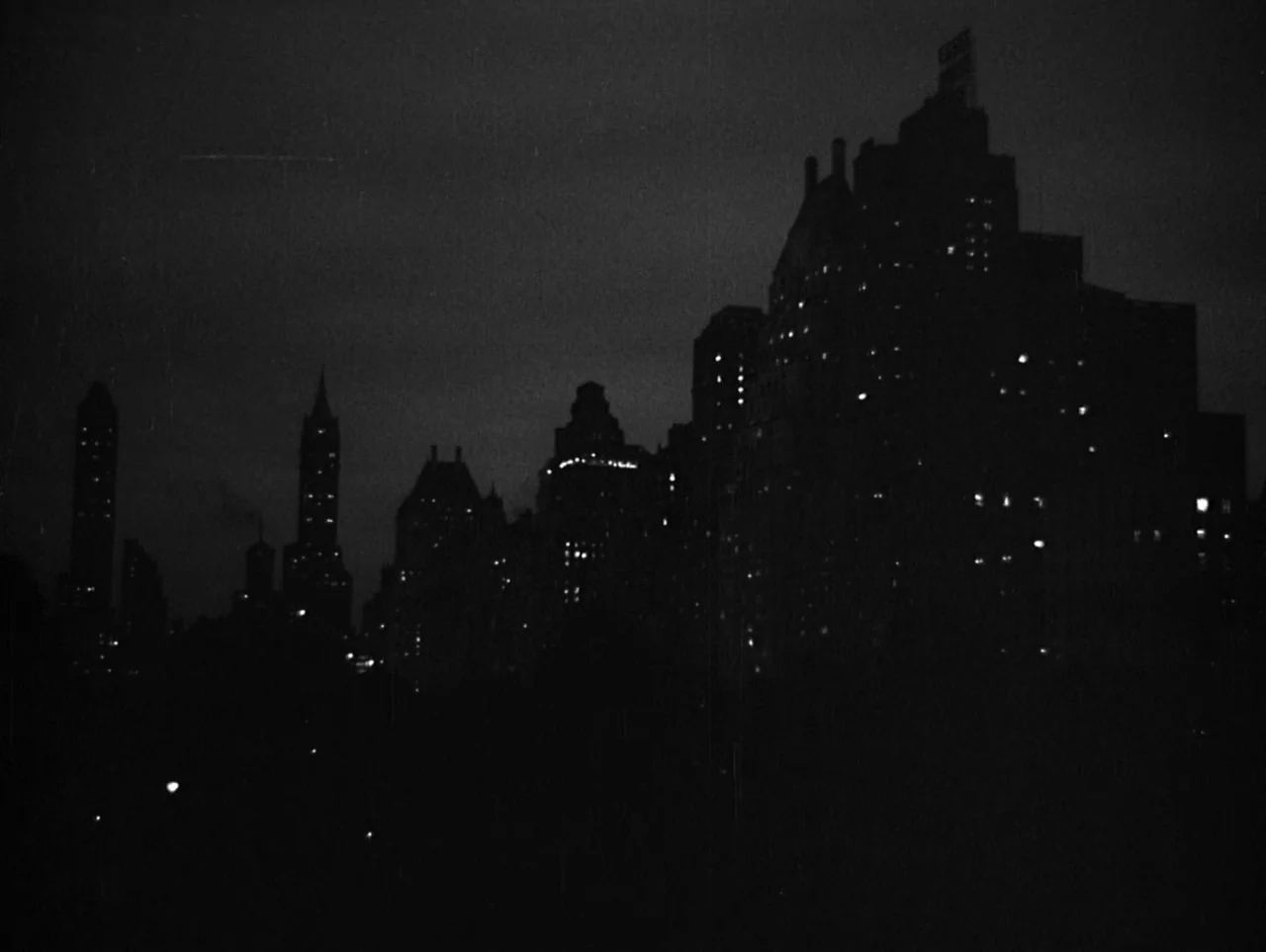 The Naked City (1948) movie still: establishing — A dark urban skyline at night features tall architectural silhouettes against a murky sky. Small…