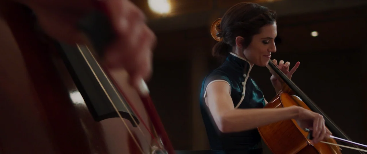 The Perfection (2018) movie still: medium — A dimly lit concert hall stage features a woman playing a cello. Another musician is partially visible…
