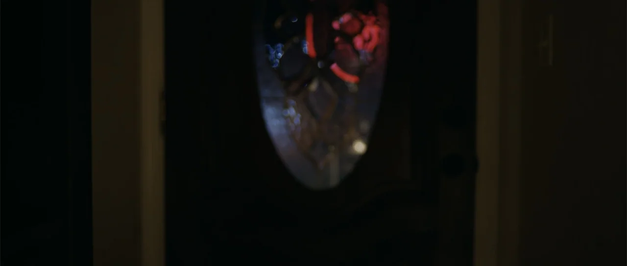 Night of the Missing (2023) movie still: medium — A dark wooden door with a central oval glass panel stands in deep shadow. Red light leaks through…