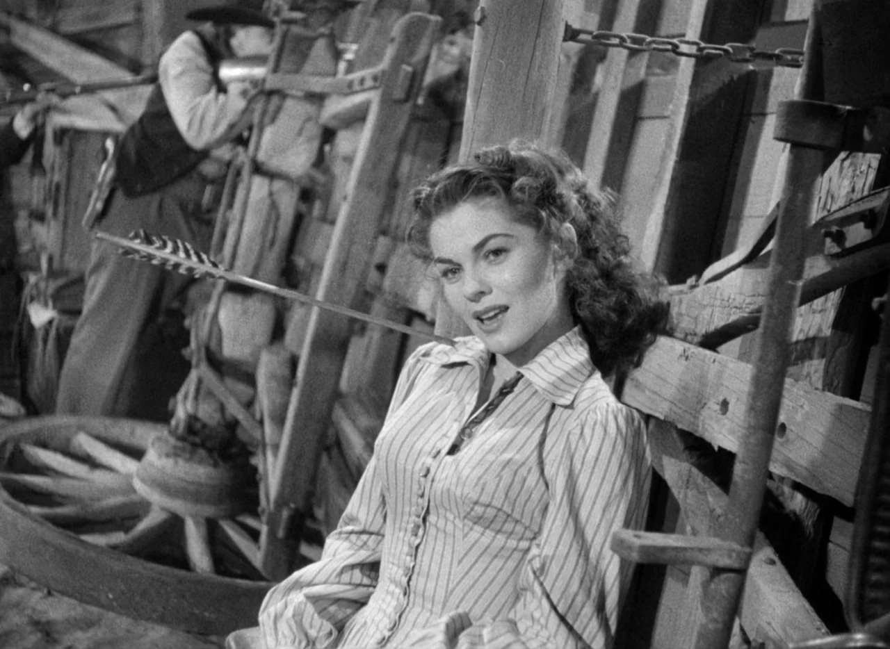 Red River (1948) movie still: medium — An interior barn setting features weathered wooden planks and vintage farm equipment. A woman leans against a…