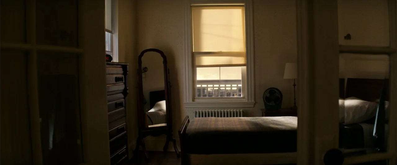 The Equalizer (2014) movie still: wide — A sparse interior bedroom features wallpaper, a single bed, and a standing wooden mirror. The room is…
