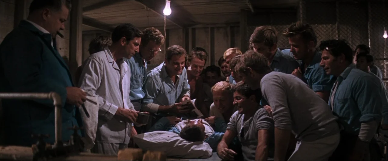 Cool Hand Luke (1967) movie still: medium — A dimly lit room with industrial walls and men clustered around a central bed. The men look down at a…