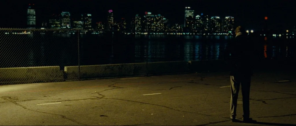 Shame (2011) movie still: wide — A dark paved waterfront area is separated from the water by a chain-link fence. A lone man stands in the foreground…