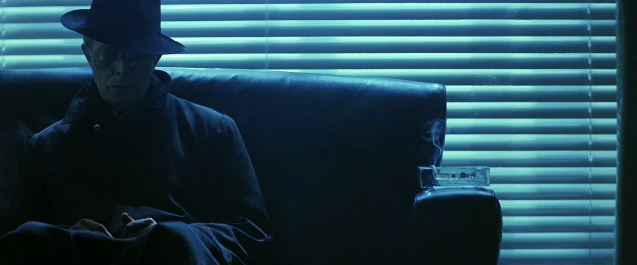 The Hunger (1983) movie still: medium — A man sits on a dark leather couch in a room with blue-tinted windows. He wears a fedora and trench coat…