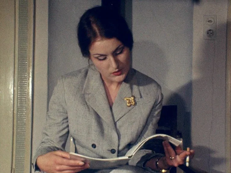 Der Bomberpilot (1970) movie still: medium — A woman sits in a room with a light-colored wall and electrical outlet. She holds an open book and a…