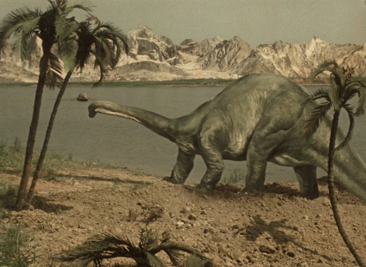 A Journey to the Beginning of Time (1955) movie still: wide — A prehistoric lake landscape features a large sauropod moving along a sandy, rocky…