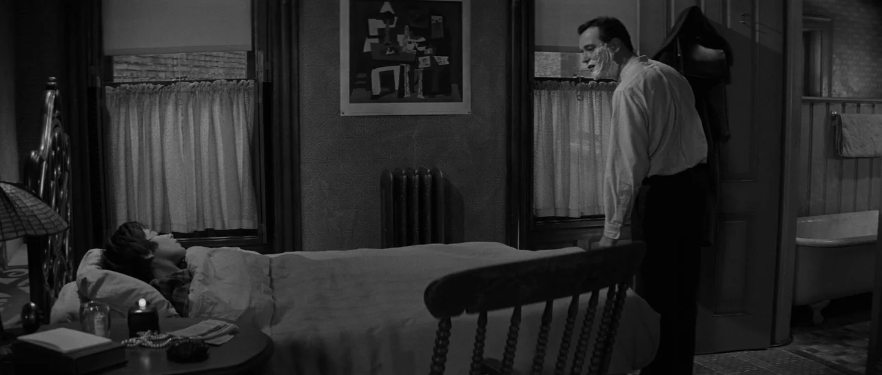 The Apartment (1960) movie still: medium — A furnished apartment with lace window curtains, a radiator, and a wall painting. A man with shaving…