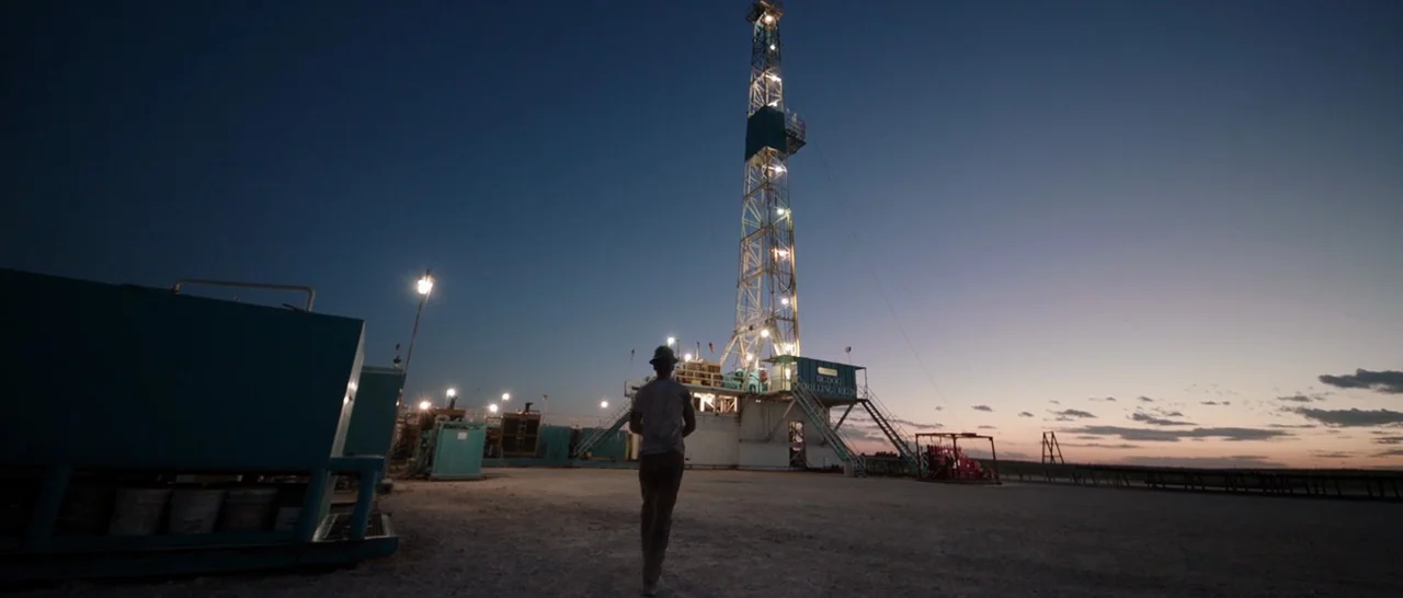 Song to Song (2017) movie still: wide — An open gravel yard surrounds a large illuminated industrial drilling rig at twilight. A man walks away from…