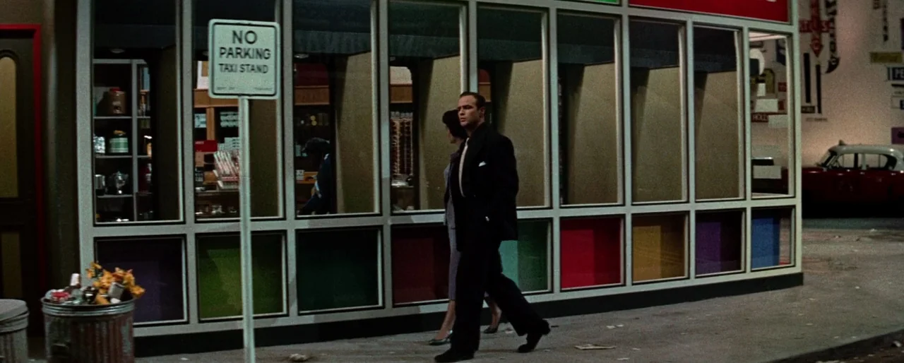 Guys and Dolls (1955) movie still: fullbody — A nighttime city sidewalk passes in front of a building with large glass windows. A man in a suit…