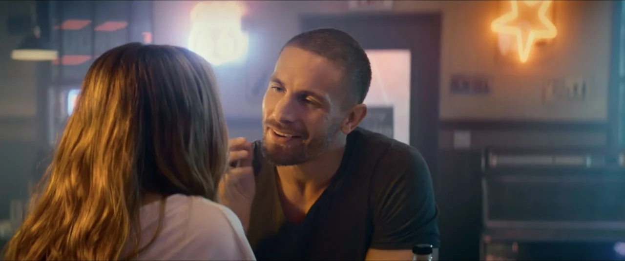 Man with No Past (2025) movie still: medium — A dimly lit eatery features neon wall signage including a star and route marker. A man and a woman sit…