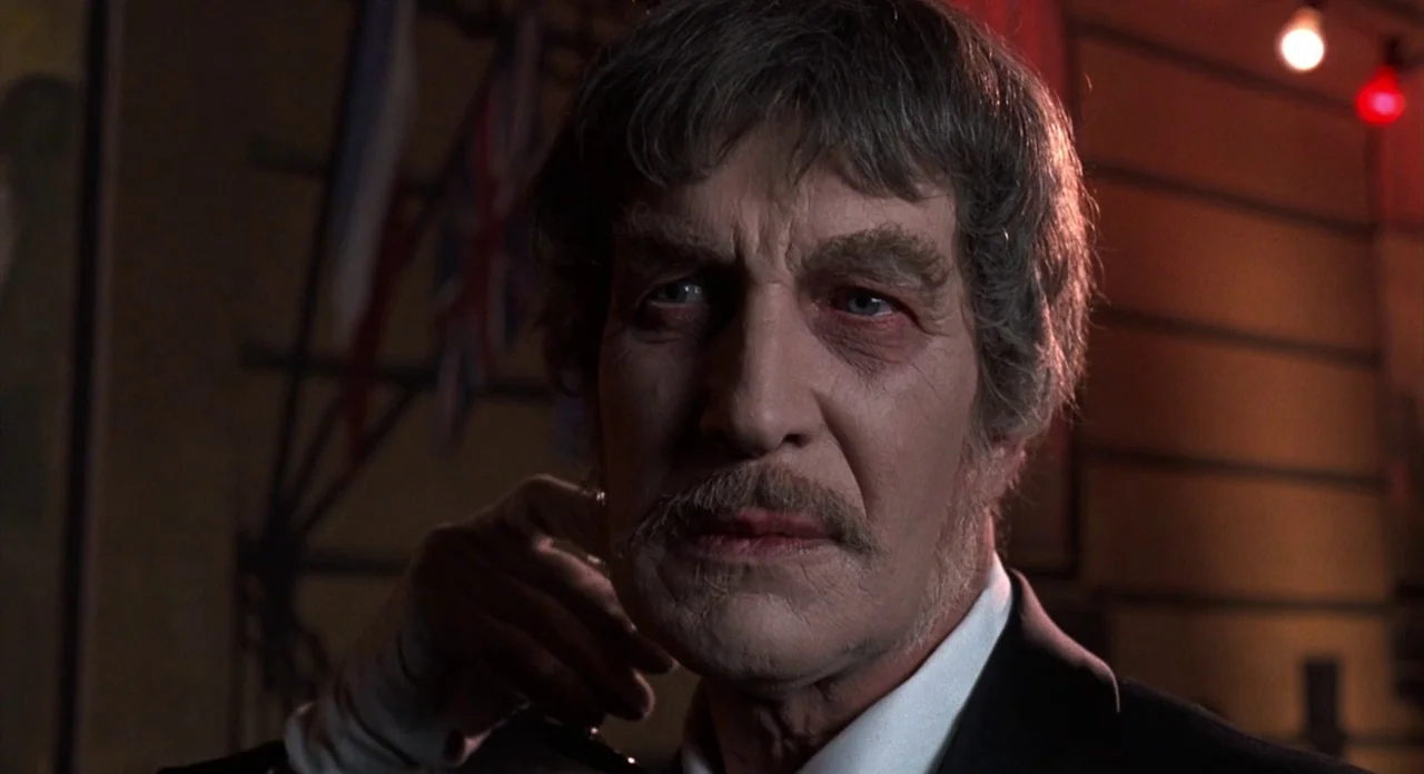 The Abominible Dr. Phibes (1972) movie still: closeup — A dimly lit indoor space featuring a wall with a British flag hanging partially. An elderly…