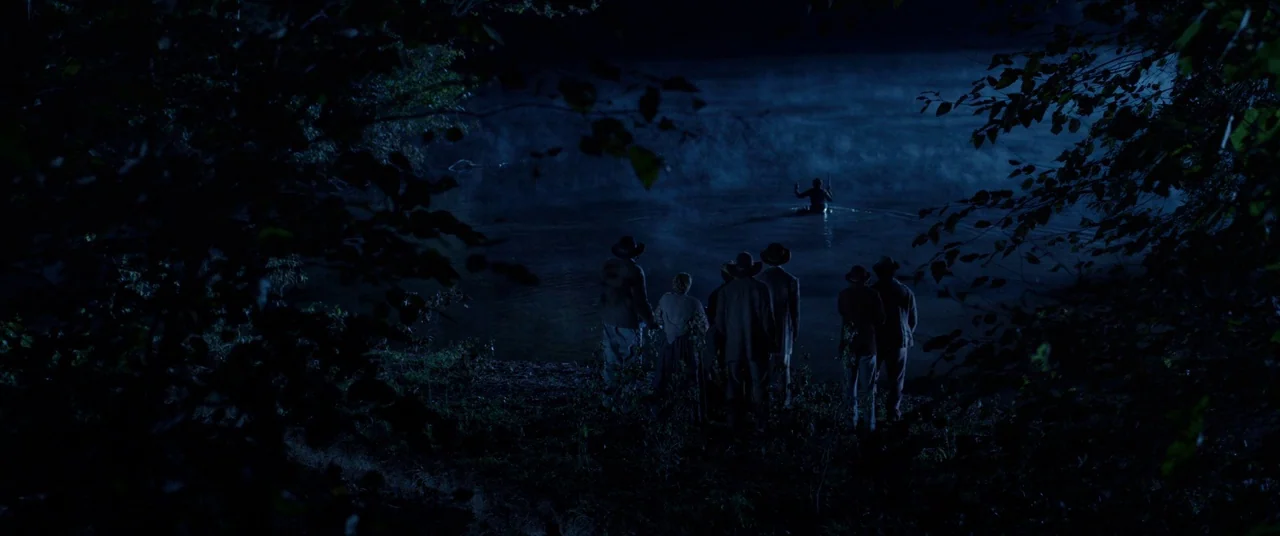 Harriet movie still: wide — A group of people standing on a dark riverbank at night under moonlight. They face away from the camera toward a lone…