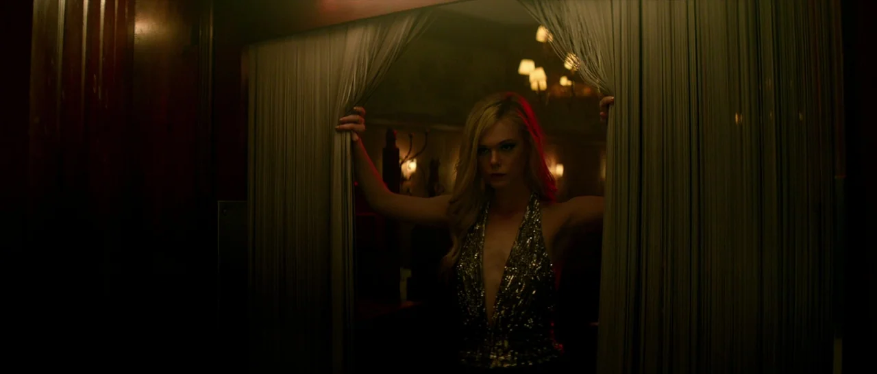 The Neon Demon (2016) movie still: medium — A dimly lit hotel room interior features dark wood paneling and soft warm lighting. A woman stands…