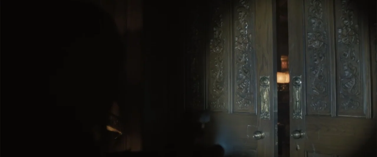 Abigail (2024) movie still: medium — A dimly lit interior space features ornate, dark wooden double doors with brass hardware. A person stands with…