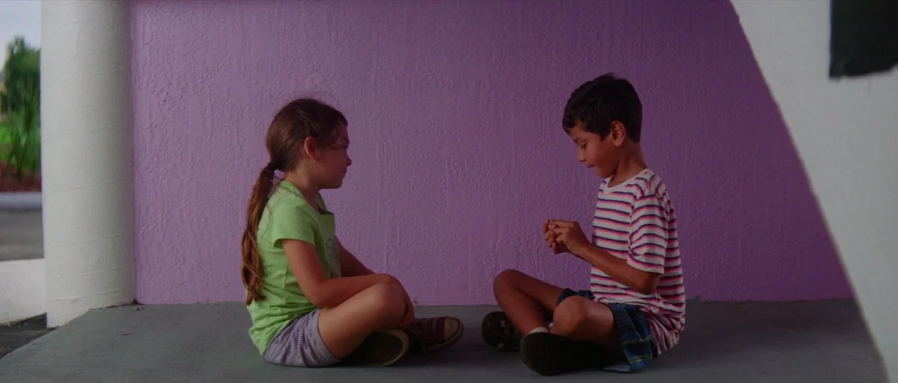 The Florida Project (2017) movie still: fullbody — Two children sit on a concrete floor against a vibrant purple exterior wall. They face each other…