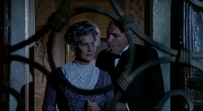 Lisa and The Devil (1973) movie still: medium — A dimly lit hallway features a decorative wrought iron gate in the foreground. A woman and a man in…