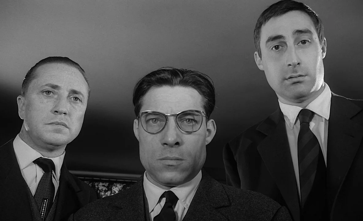 The Trial (1962) movie still: medium — Three men in suits stand closely together in an indoor setting with a plain background. They stare downward…