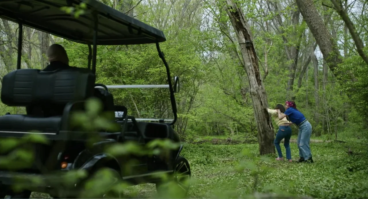 Turtles All the Way Down (2024) movie still: wide — A dense green forest area features a parked black golf cart in the foreground. Two women in…