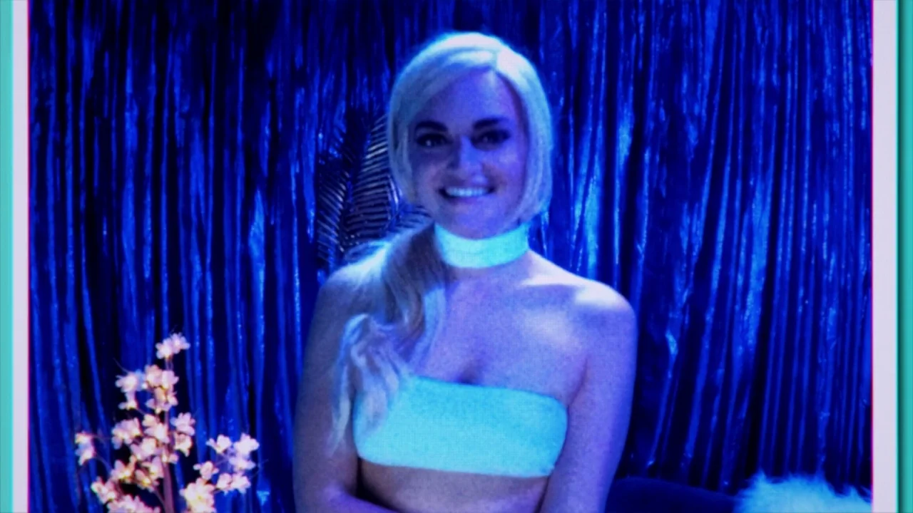Cam (2018) movie still: medium — A woman stands in front of a blue reflective curtain in a studio setting. She looks toward the camera while wearing…
