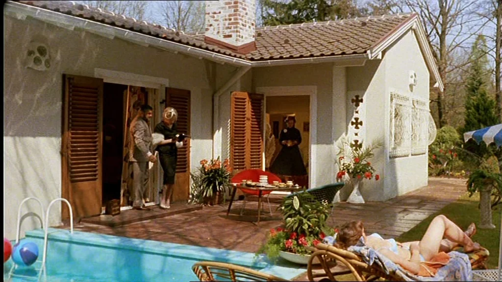 Lola (1981) movie still: wide — A cream-colored house with wooden shutters features a small patio beside a swimming pool. Three people stand near…