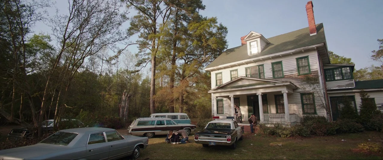 The Conjuring (2013) movie still: wide — A two-story white farmhouse with green shutters sits behind several vintage 1970s vehicles. A group of…