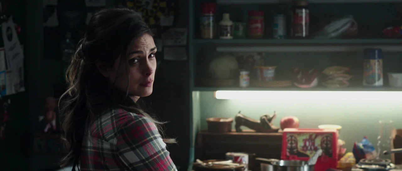 Deadpool (2016) movie still: medium — A dimly lit room features shelves filled with canned goods and a small red lunchbox. A woman turns her head to…