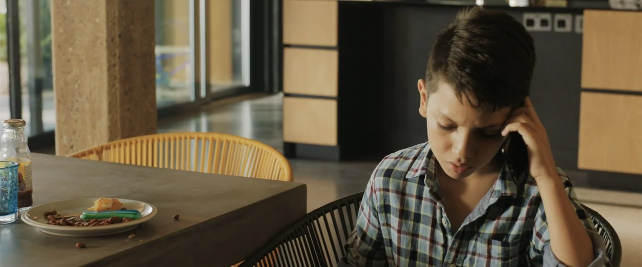 The 355 (2022) movie still: medium — A modern interior space with wood accents and a concrete dining table surface. A boy sits at the table using a…