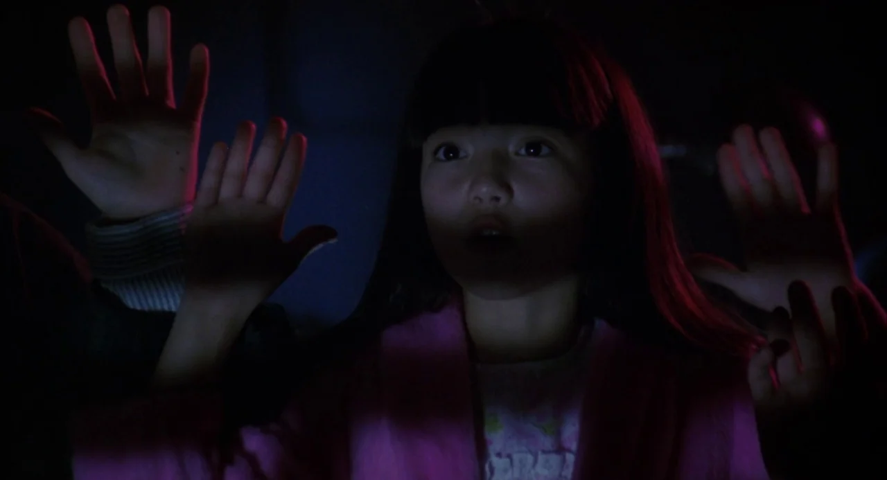 Night of the Comet movie still: medium — A dark, interior setting where only the face and hands of a young girl are visible. The subject presses her…
