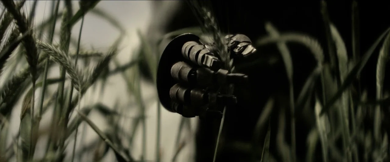 Rebel Moon – Part One: A Child of Fire (2023) movie still: closeup — A metal prosthetic hand reaches into a patch of tall green wheat stalks. The…
