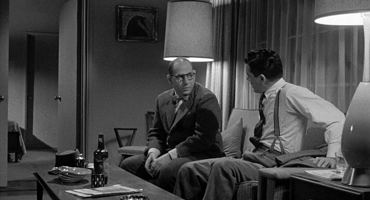 Murder By Contract (1958) movie still: medium — A mid-century modern living room features a sofa, a coffee table, and large lamps. Two men sit on…