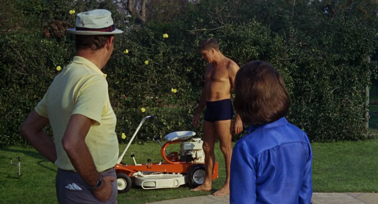 The Swimmer (1968) movie still: medium — A manicured lawn surrounds a small orange riding mower next to dense green hedges. Three people stand in a…