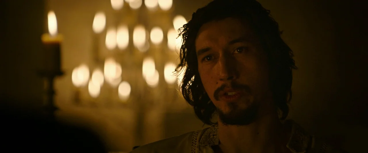 The Last Duel (2021) movie still: closeup — A dimly lit medieval stone room with an out-of-focus chandelier of burning candles behind. A man with…