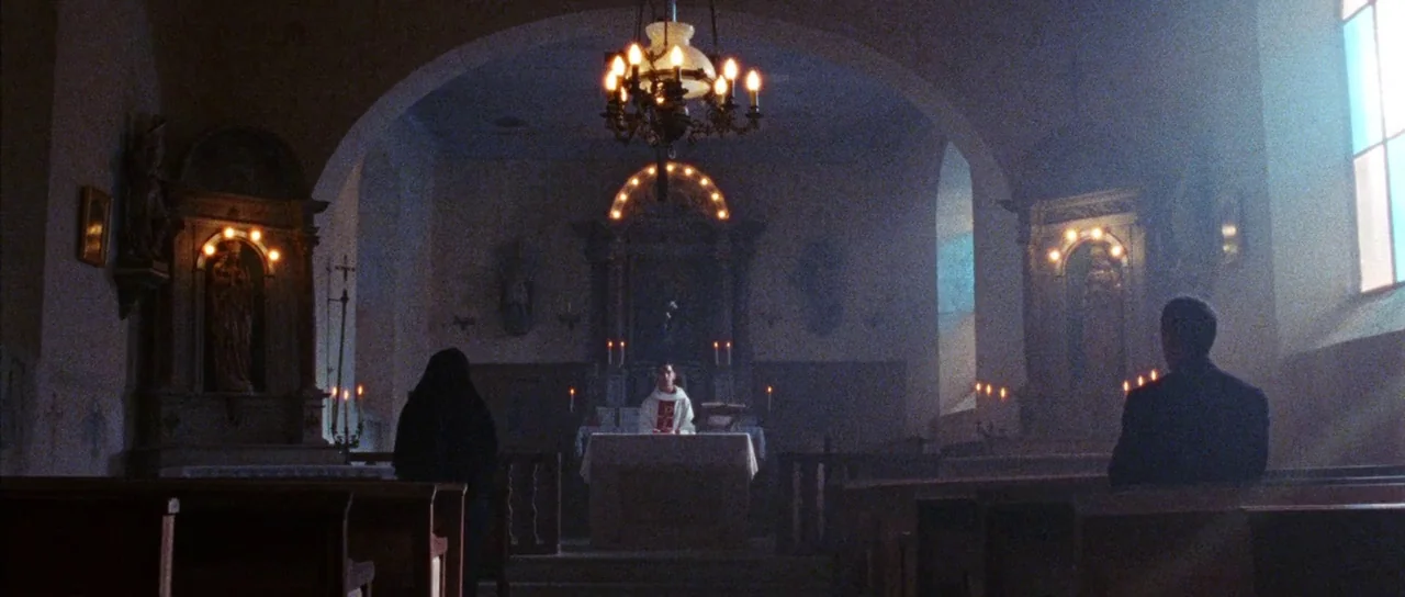 Alléluia (2014) movie still: wide — A dimly lit church interior features stone arches and wooden pews in soft fog. A priest stands at the central…