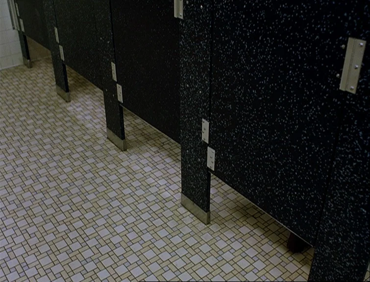 Elephant (2003) movie still: wide — A row of dark speckled partitions divides several bathroom stalls in a sterile facility. The floor is covered in…