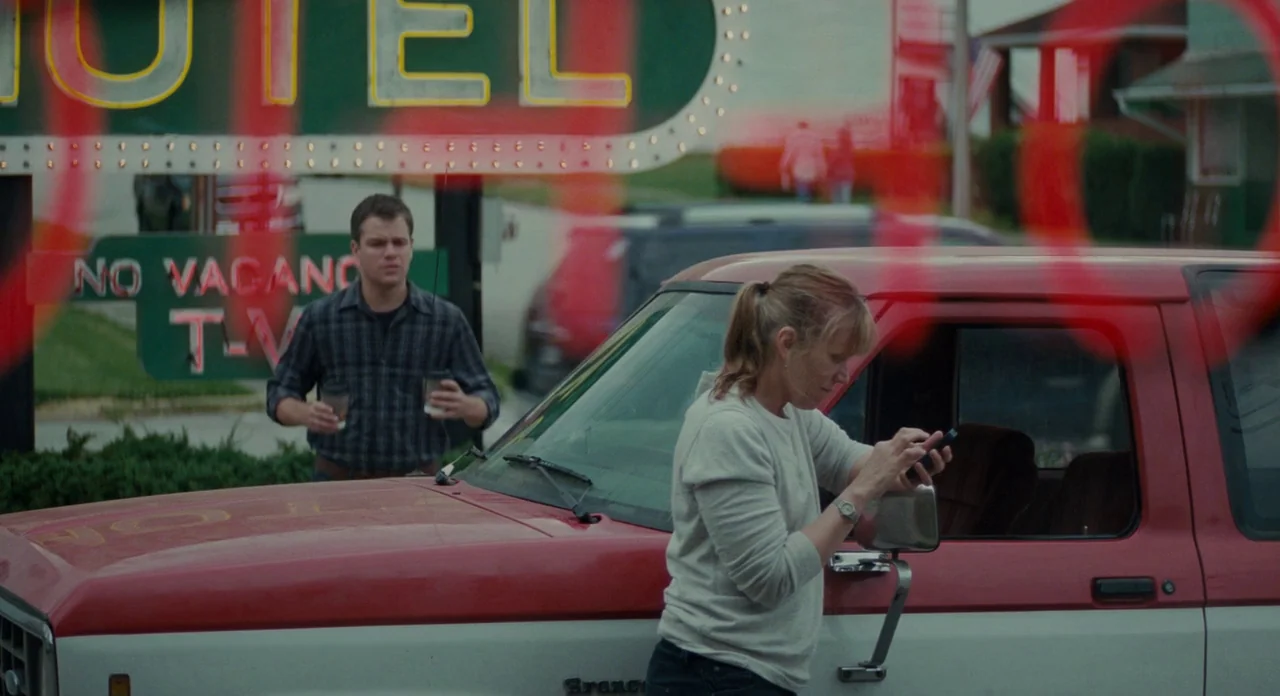 Promised Land (2012) movie still: medium — A suburban motel parking lot features a vintage red and white Ford Bronco parked nearby. A man approaches…