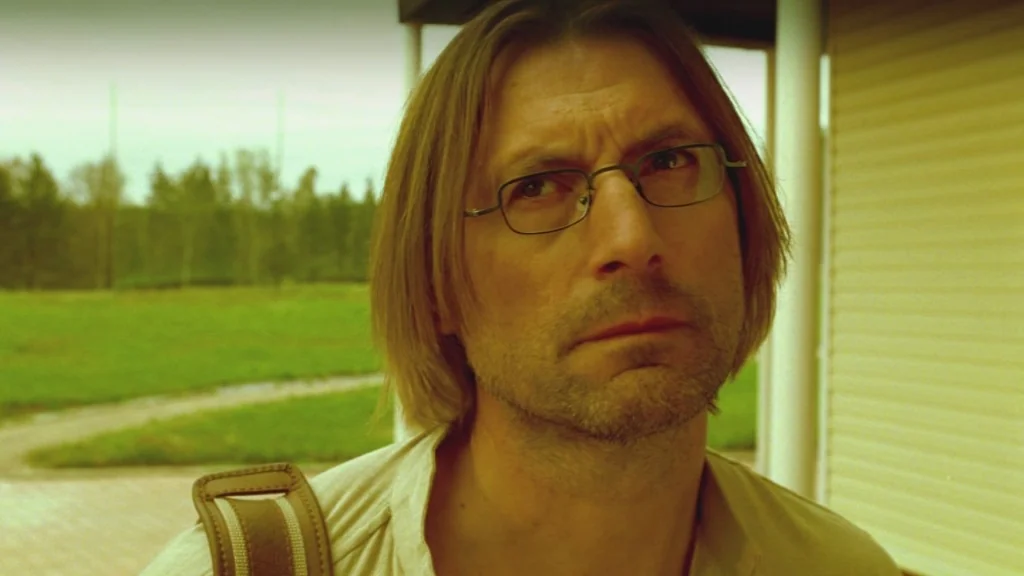 The Ugly Swans (2006) movie still: closeup — A man stands on a porch with green fields and trees in the distance. The man looks toward the side…