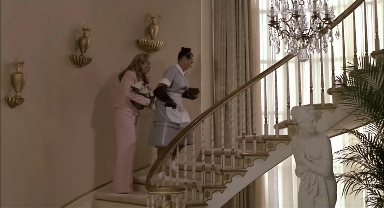 She-Devil (1989) movie still: fullbody — A grand residential staircase features a white stone statue and an ornate crystal chandelier. Two women…
