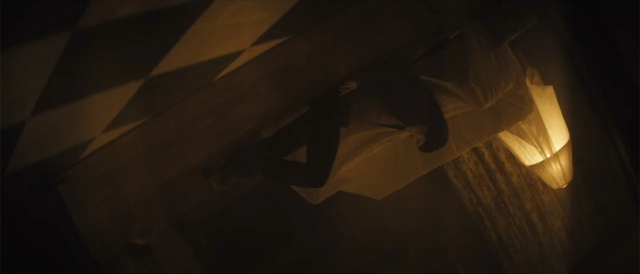 All Fun and Games (2023) movie still: medium — A dimly lit room features a person reclining on a bed beside a lamp. The camera is rotated at a dutch…