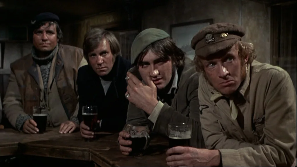 Straw Dogs (1971) movie still: medium — A dimly lit room with wood paneled walls and a worn bar counter. Four men sit side by side holding glasses…