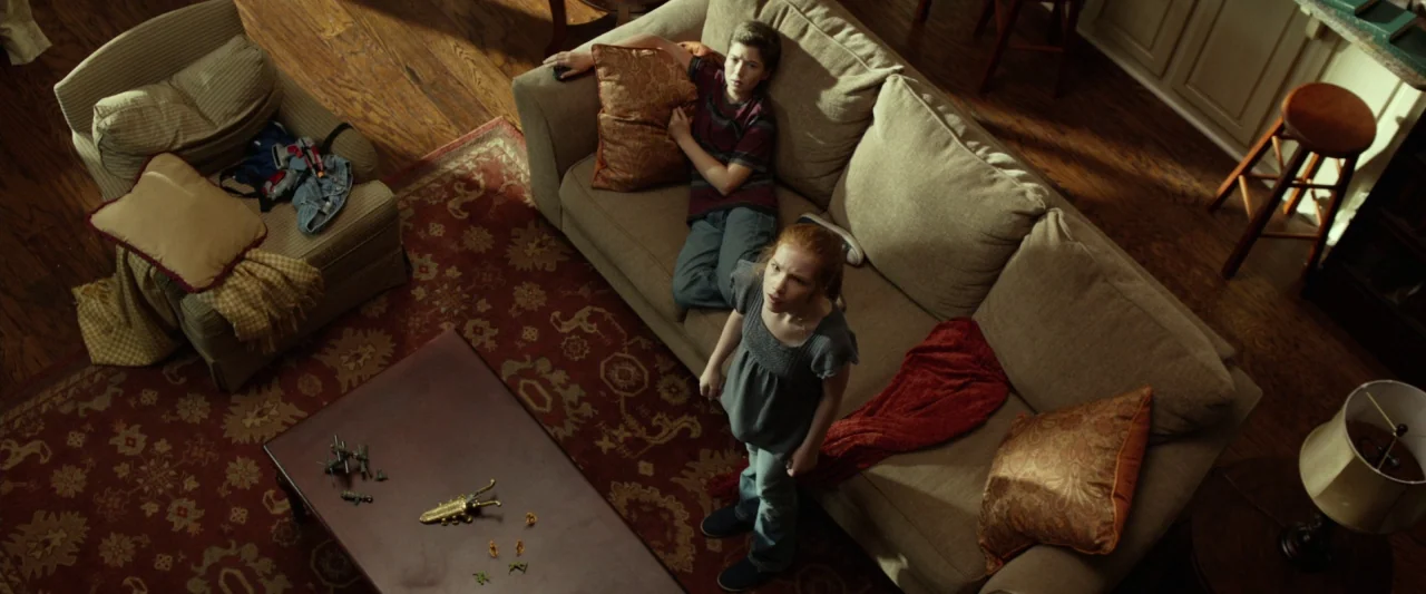 Oculus (2013) movie still: wide — A living room features a tan sectional couch set upon an ornate red rug. A boy rests on the sofa while a young…