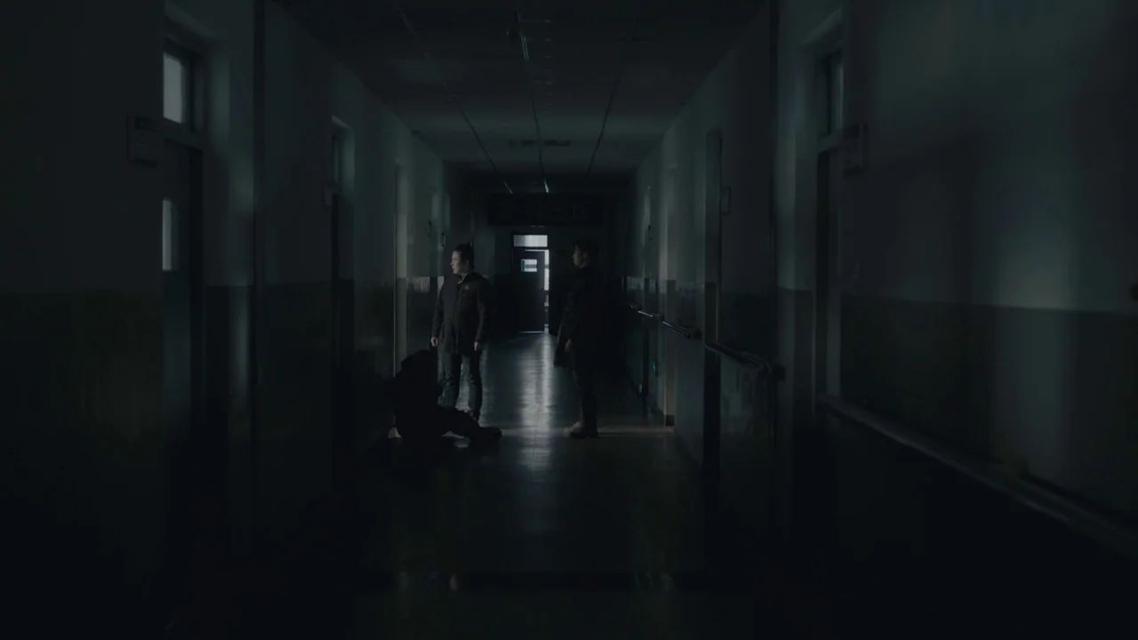 An Elephant Sitting Still (2018) movie still: wide — A dimly lit hospital corridor stretches into darkness with doors lining both walls. Three men…