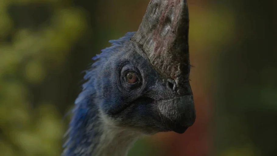 Prehistoric Planet (2022) movie still: closeup — A close-up of a blue feathered dinosaur head with a prominent bony crest. The subject looks forward…