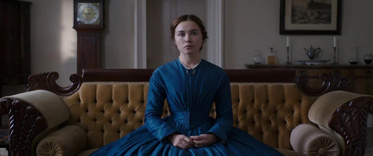 Lady Macbeth (2016) movie still: medium — A woman sits centered on an ornate yellow tufted sofa in a dimly lit room. A tall grandfather clock and…