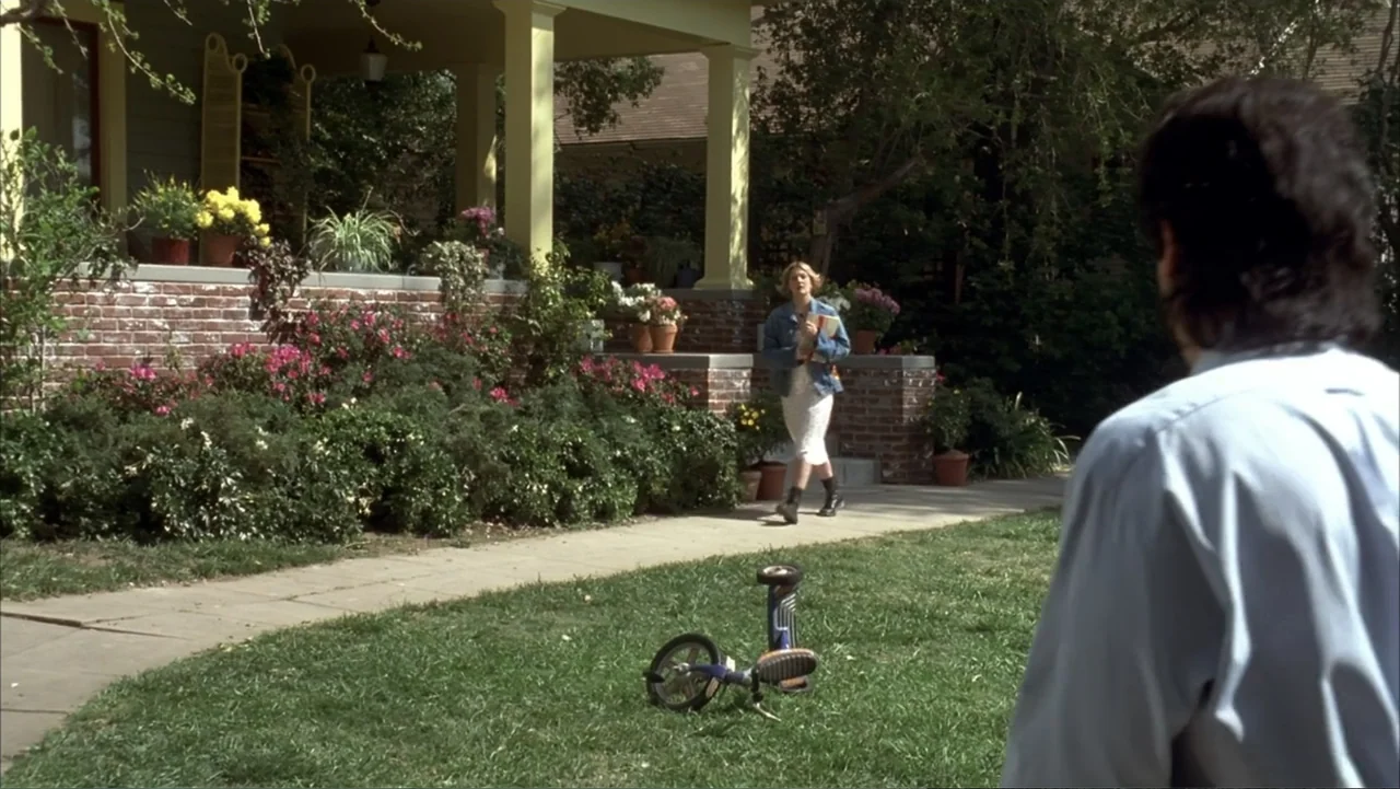 The Wedding Singer (1998) movie still: wide — A suburban house with a brick-bordered flower bed and a mowed lawn. A woman in a denim jacket walks…
