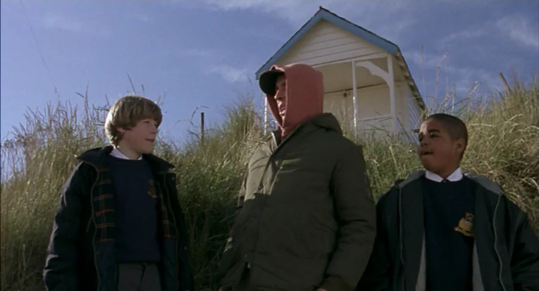 A Room For Romeo Brass (1999) movie still: medium — A grassy sand dune is occupied by three people with a small wooden structure behind. The trio…