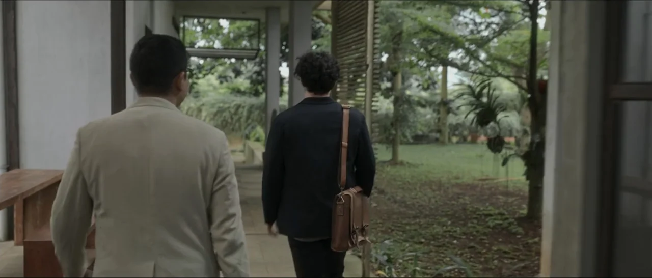 Innocent Vengeance (2023) movie still: medium — A covered walkway with concrete pillars connects to a lush green outdoor area. Two men in jackets…